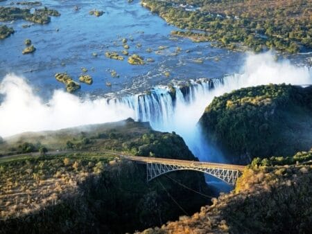 Blick auf die tosenden Victoria Fälle aus der Luft. Im Vordergrund ist die Eisenbahnbrücke zu sehen.