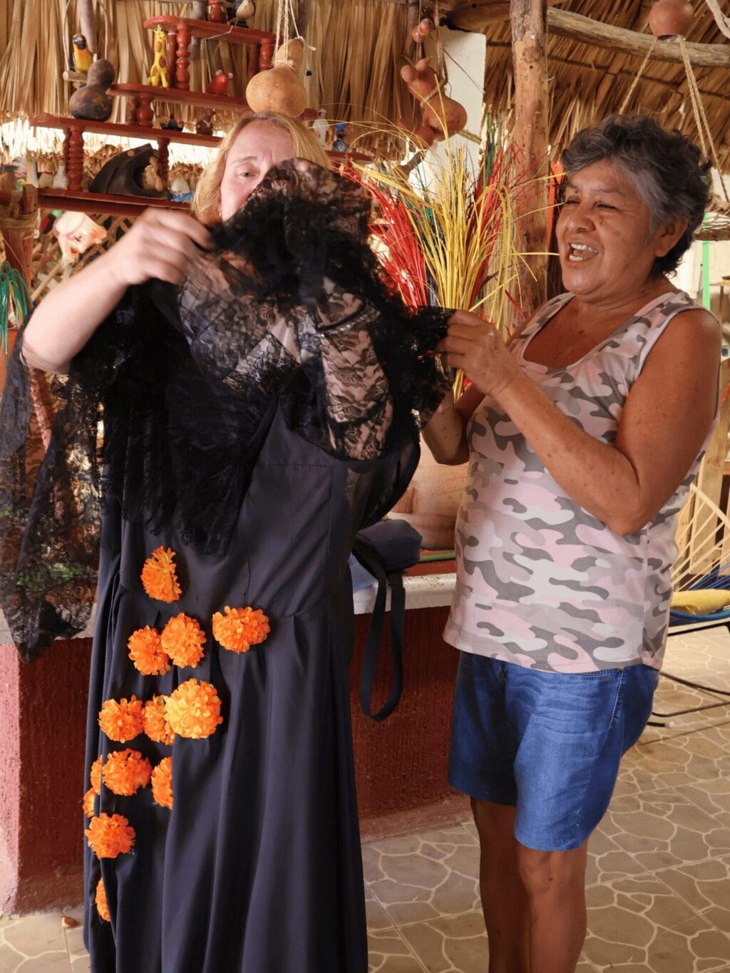 Eine mexikanische Frau mit schwarz-grauen Haaren, blauen kurzen Hosen und einem hellen, gemusterten Träger-T'Shirt hilft einer blonden Frau, ein schwarzes Kleid mit orangefarbenen Pompoms anzuziehen. Sie befinden sich in einer Halle unter einem Strohdach.